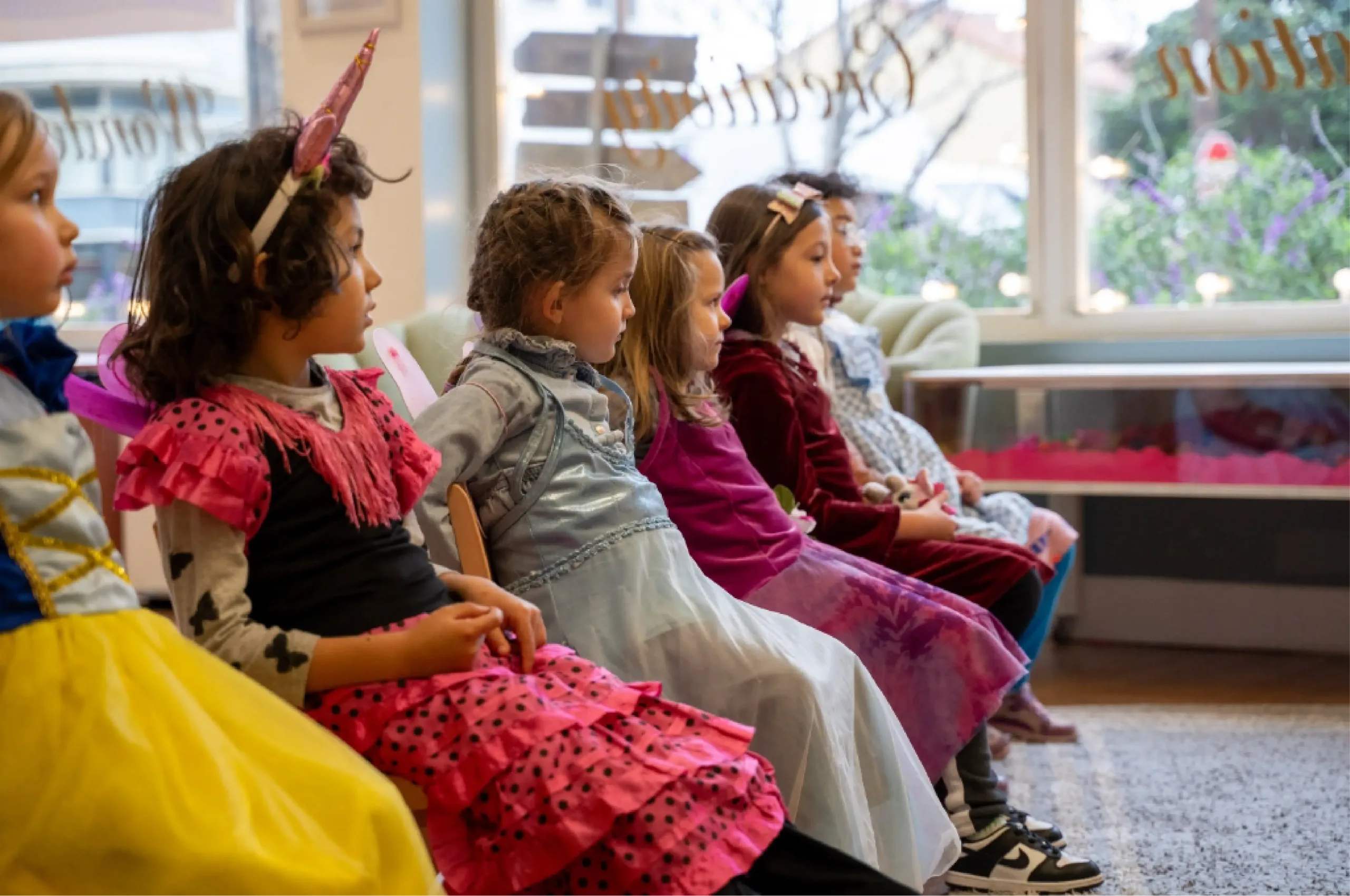 Five young children sit in a row indoors, wearing colorful costumes, facing towards the right side of the room.