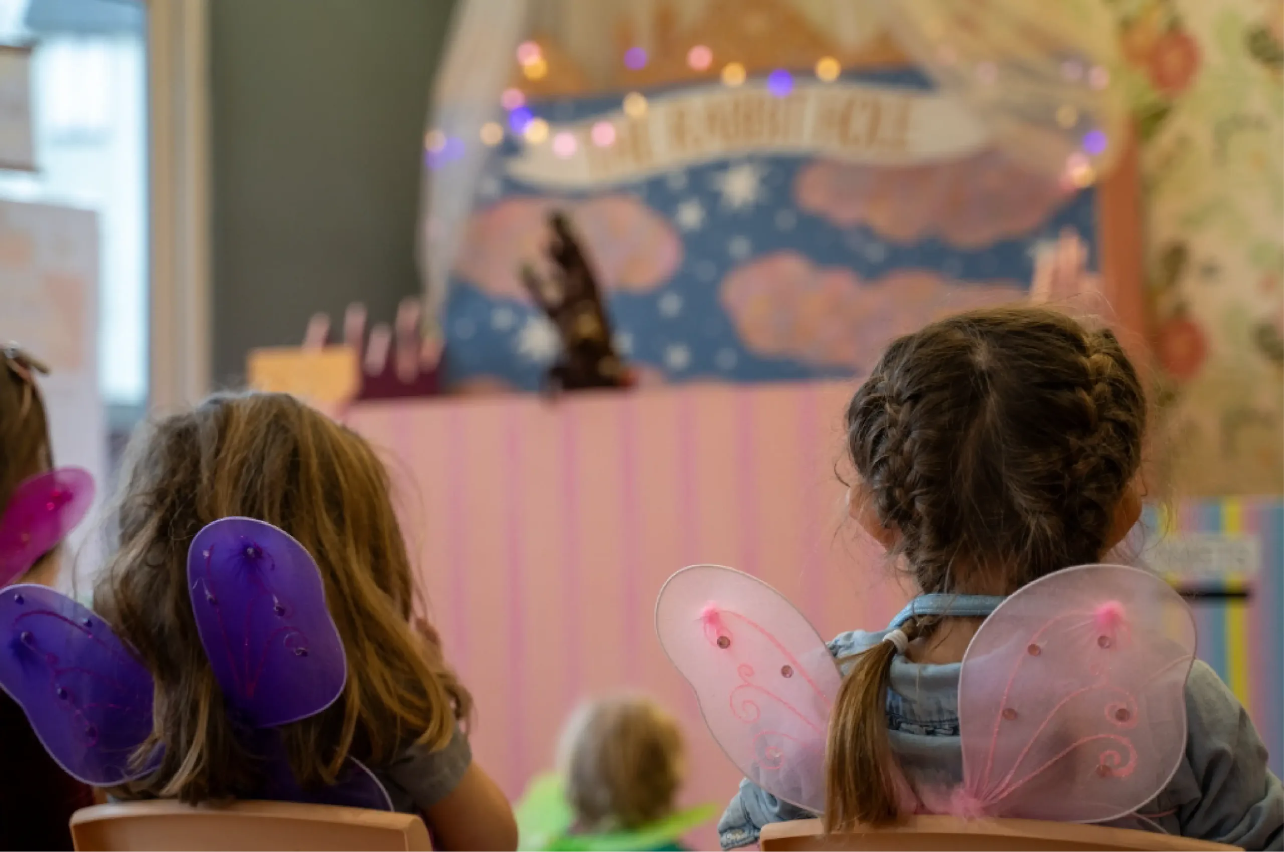 Children wearing fairy wings watch a puppet show on a decorated stage with a backdrop featuring clouds and lights.