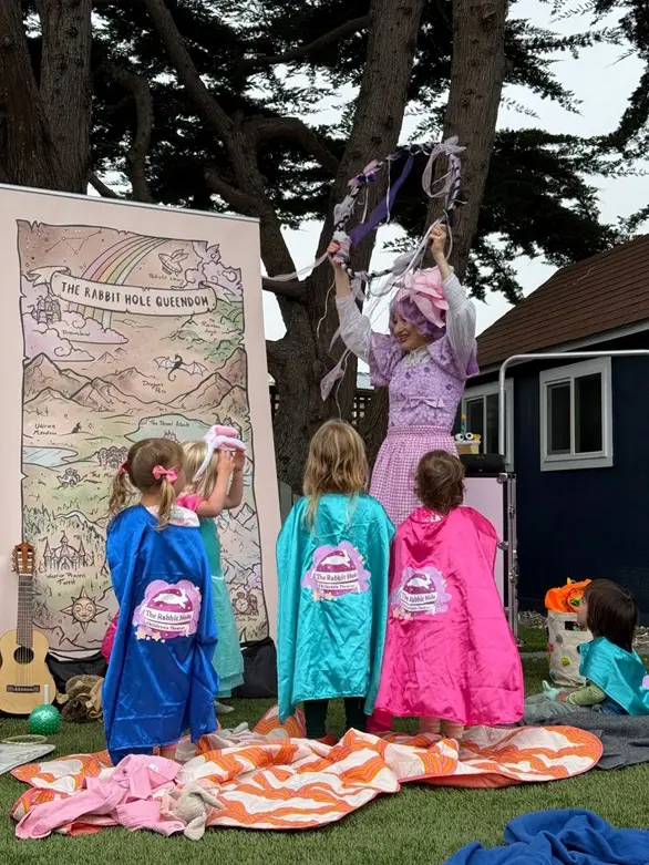 A woman in costume from The Rabbit Hole Children’s Theater stands in front of a map, performing for four young children in colorful capes outdoors, with trees and a house in the background.