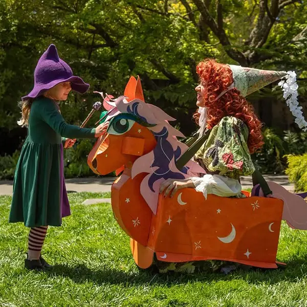 A child in a purple hat and dress holds a wand while facing an adult dressed as a fairy or witch, sitting on an orange horse costume outdoors on grass.