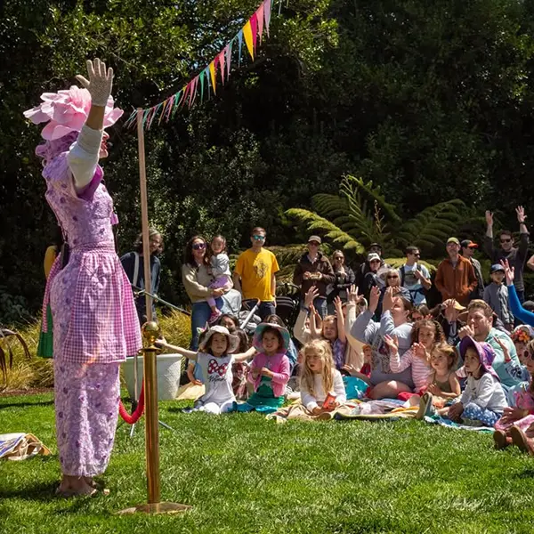 A performer in a pink costume entertaining a group of children and adults seated on grass outdoors; many audience members have raised hands.
