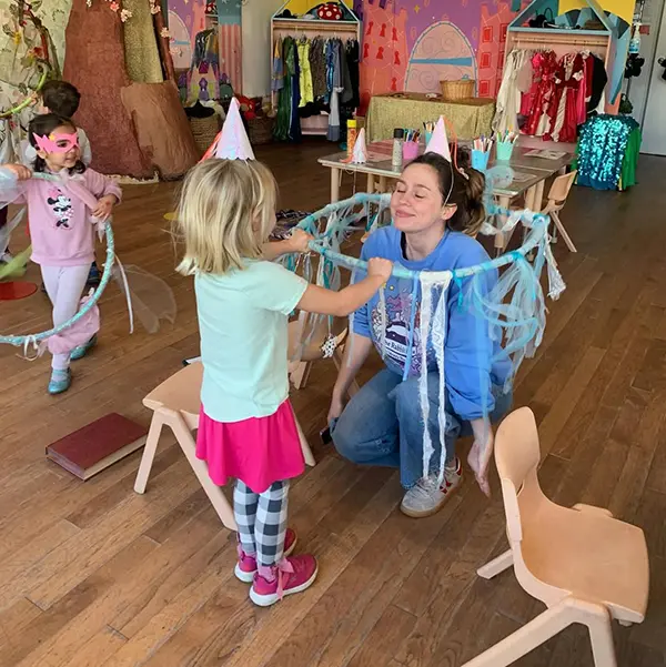 A young girl taps a kneeling woman on the shoulder with a ribbon wand in a decorated room, as other children in costumes play nearby, capturing a magical moment all about childhood imagination.