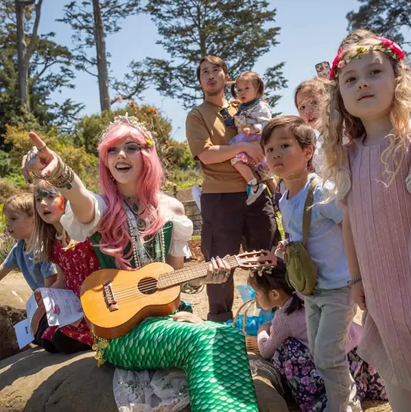 A person dressed as a mermaid with a pink wig and green tail plays a ukulele for a group of children outdoors on a sunny day.