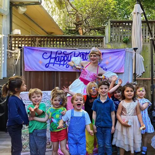 A group of children pose with a woman dressed as a princess in front of a "Happy Birthday" banner at an outdoor party.