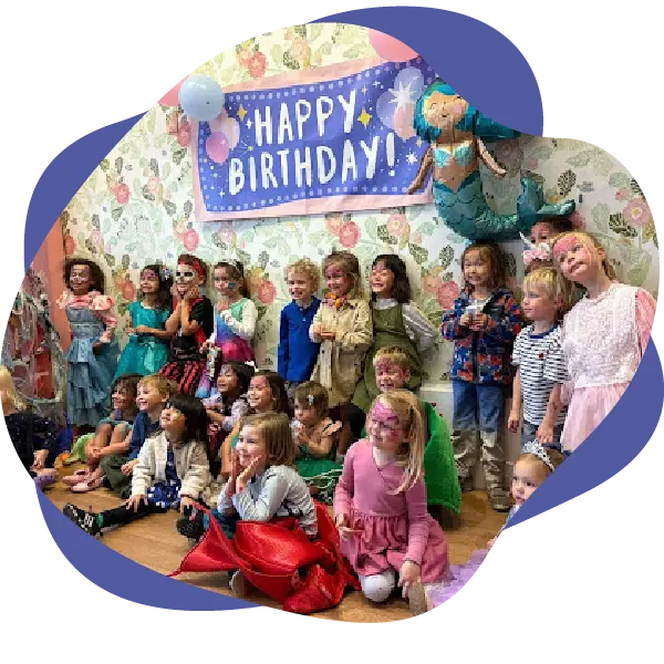 A group of children pose together at a birthday party in front of a "Happy Birthday!" banner with balloons and decorations on the wall at The Rabbit Hole Children’s Theater.