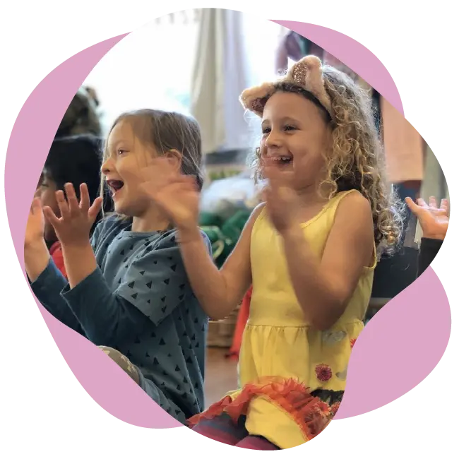 Two young children sit side by side, smiling and clapping their hands. One wears a blue shirt and the other, in a yellow dress with animal ear headbands, brings the magic of The Rabbit Hole Children’s Theater to life.