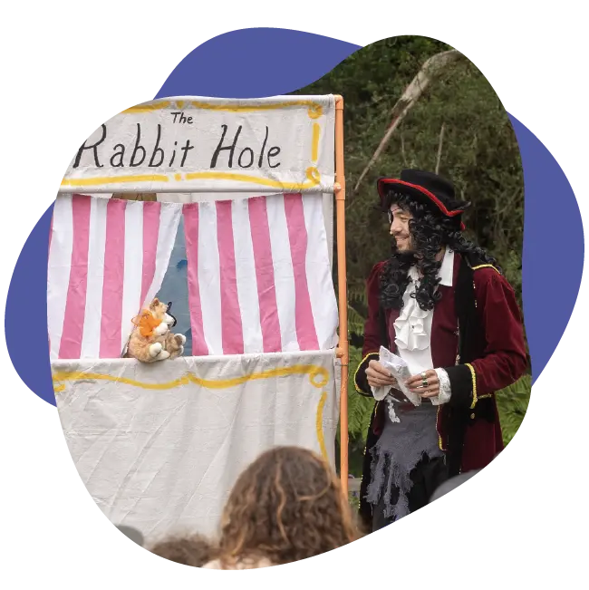 A person dressed as a pirate stands next to a puppet show booth labeled "The Rabbit Hole Children’s Theater," with a puppet visible behind the striped curtain.