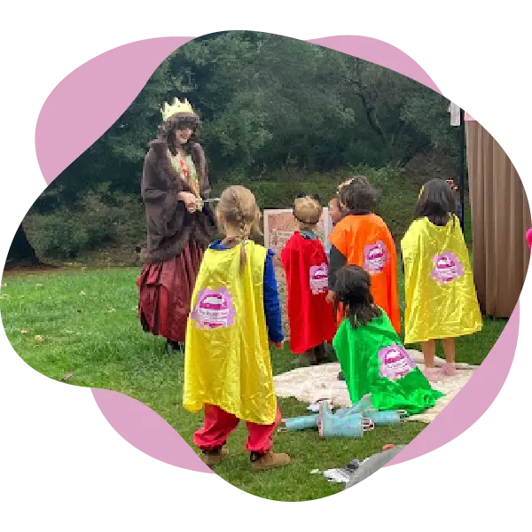 An adult in a crown and cape stands outdoors facing a group of children in colorful capes, with trees and grass in the background—an imaginative scene brought to life by The Rabbit Hole Children’s Theater.