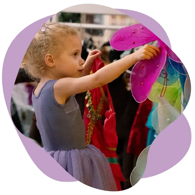 A young child in a purple dress reaches toward colorful butterfly wings hanging on a rack, discovering creativity through Studio Programs.