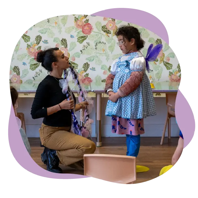 An adult kneels and talks to a child wearing a costume with wings in a classroom decorated with floral wallpaper, highlighting the creativity fostered through Studio Programs.