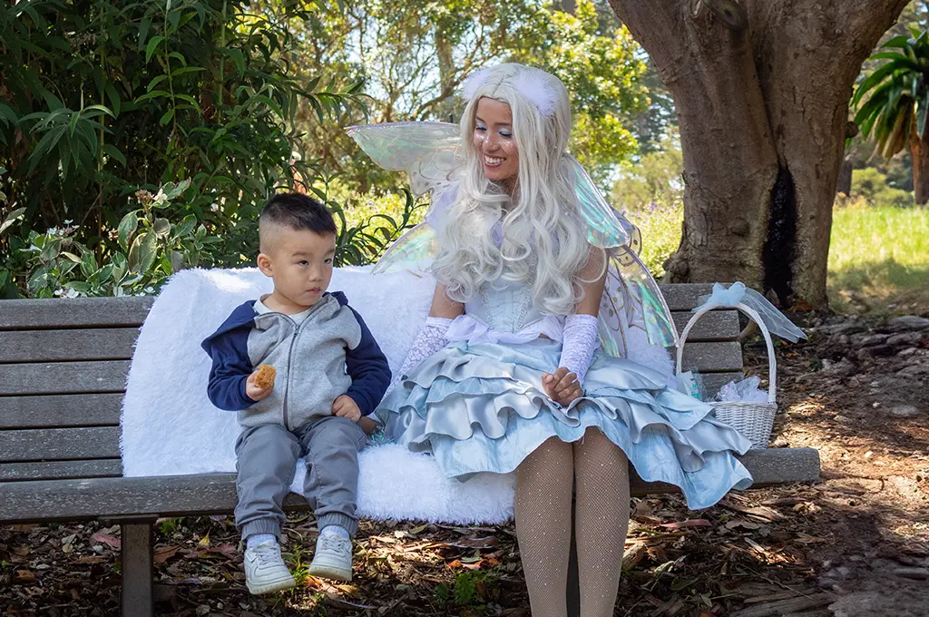 A young boy holding a snack sits on a park bench next to a woman dressed as a fairy—white hair, wings, and a blue costume—like those from Storyteller Birthday Parties. They are outdoors near trees and greenery.