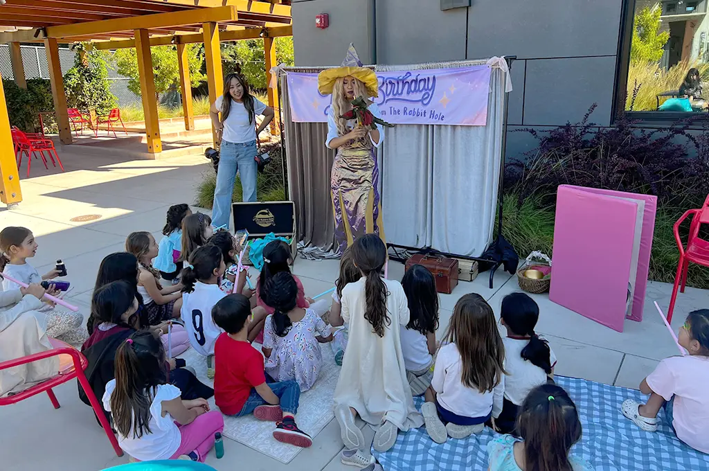 A woman in a wizard costume reads to a group of children seated outdoors, with a banner and props set up behind her—perfect inspiration for magical Storyteller Birthday Parties.