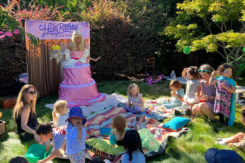 A group of children sit on blankets outdoors around a woman dressed as a princess in front of a "Happy Birthday" sign, enjoying enchanting Storyteller Birthday Parties at a garden celebration.