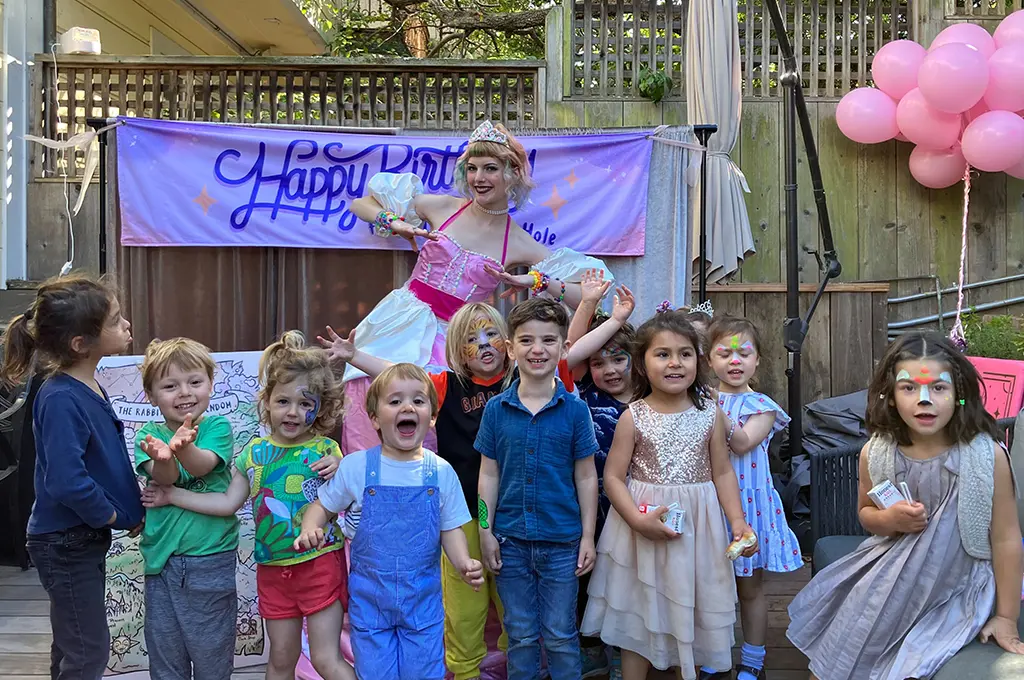 A group of children with face paint pose in front of a "Happy Birthday" banner and a princess from Storyteller Birthday Parties at an outdoor celebration. Pink balloons are on the right.