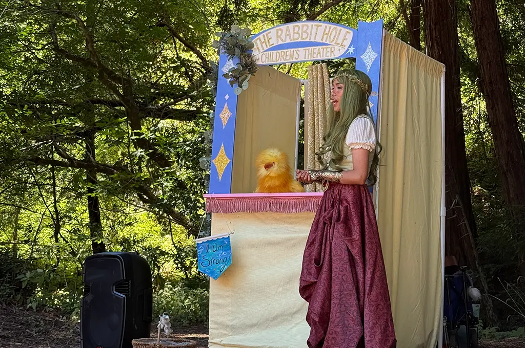 A woman in costume stands beside a puppet theater labeled "The Rabbit Hole Children's Theater," interacting with a yellow puppet outdoors—perfect for magical Storyteller Birthday Parties.